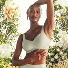 Lifestyle image of the Beyond Bra in Aloe on a model with one arm raised above her head and a background of green and white florals. 