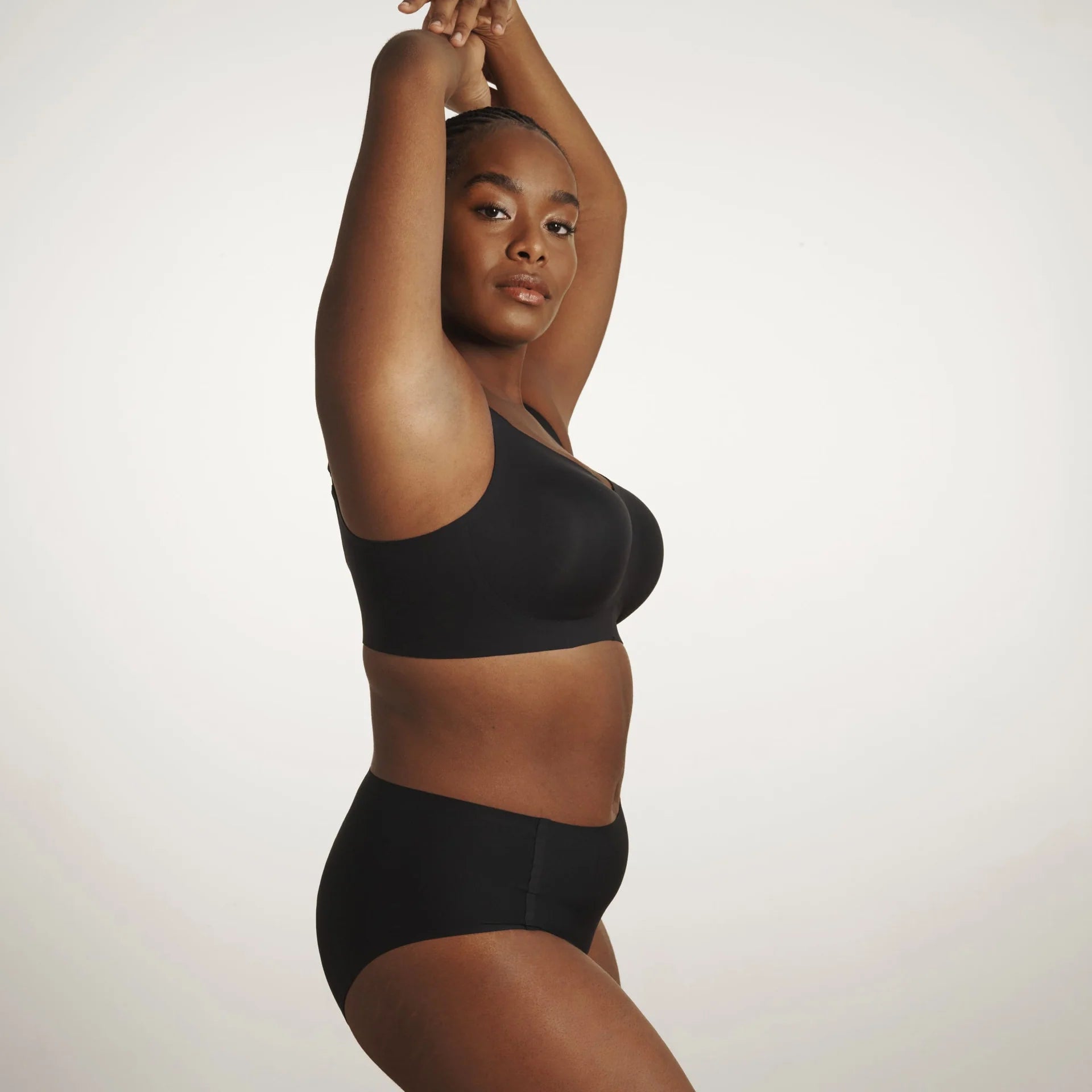 The Beyond Bra in Black, a wirefree bra shown on a model with her arm up to see the side view of the band and how it fits underneath your armpit. 
