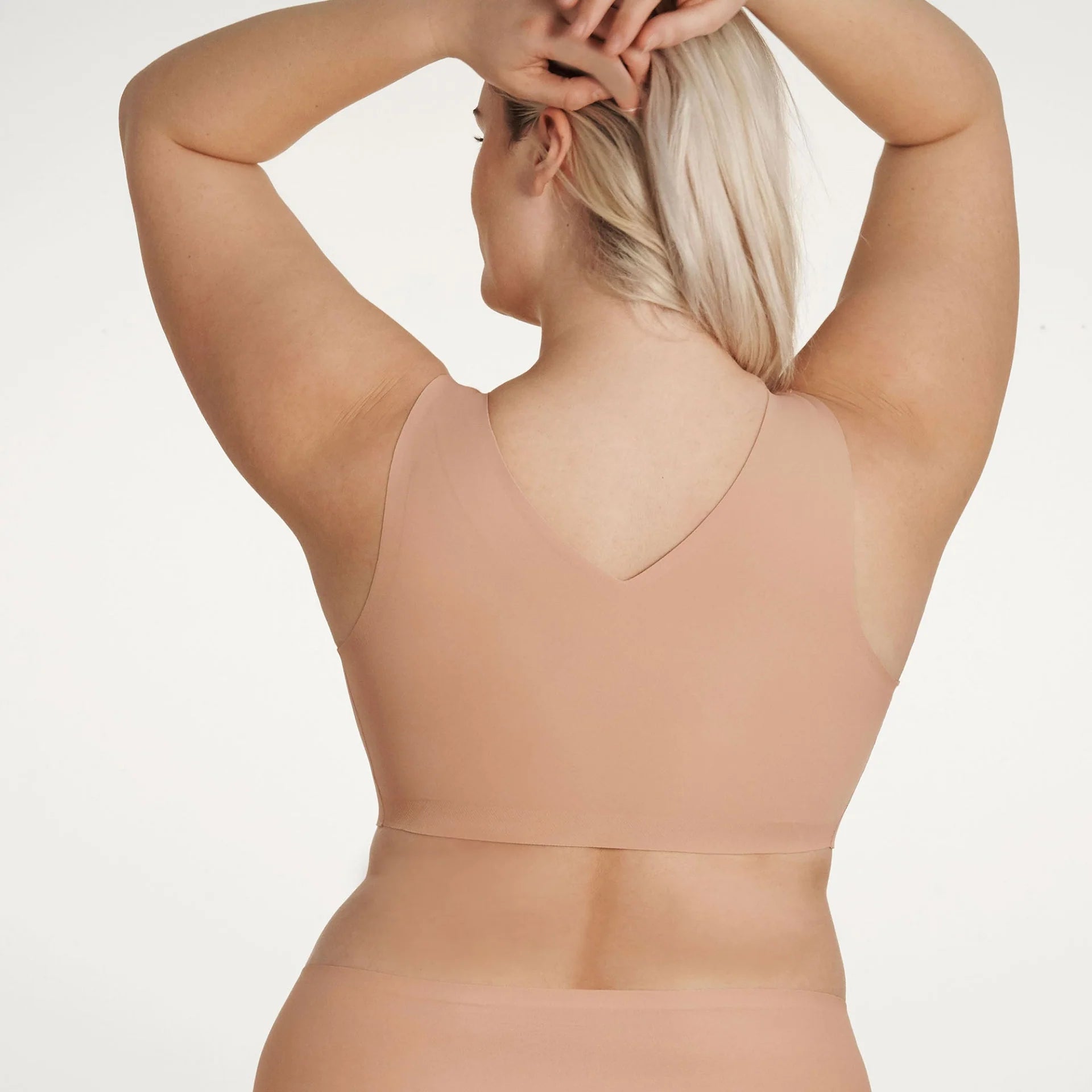 Woman wearing the Evelyn & Bobbie Defy bra in Himalayan Salt, light pinkish beige no wire pullover bra, against a white background, back view. 