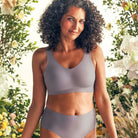 Woman wearing the Evelyn Bobbie Defy Bra in London Fog, light grey no wire bra, with a greenery and floral background. 