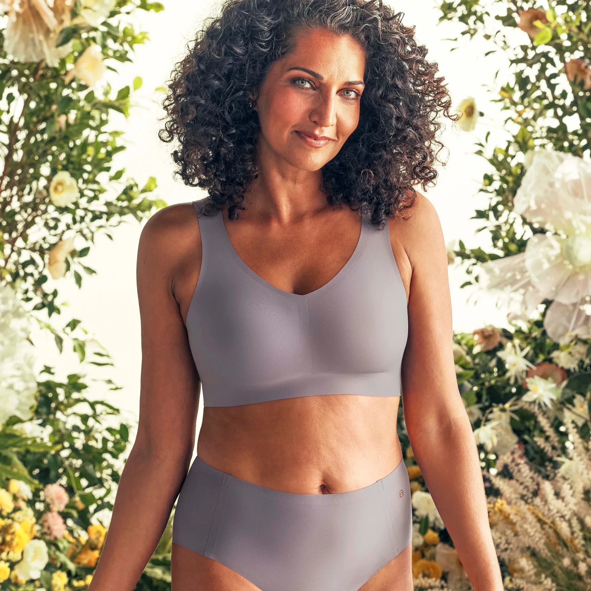 Woman wearing the Evelyn Bobbie Defy Bra in London Fog, light grey no wire bra, with a greenery and floral background. 
