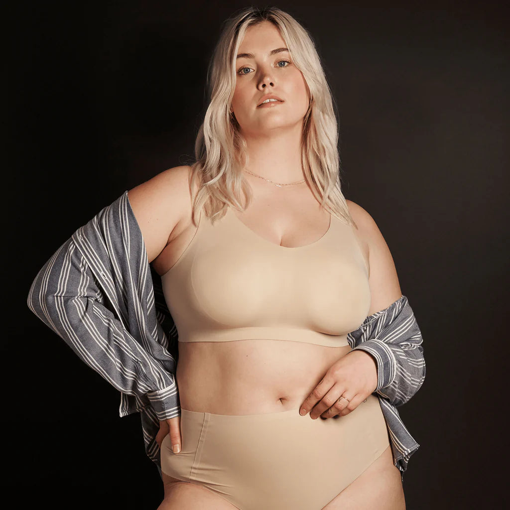 A woman wearing the Evelyn & Bobbie Defy Bra in Sand, light beige no wire bra, on a black background, with a striped shirt on her arms. 