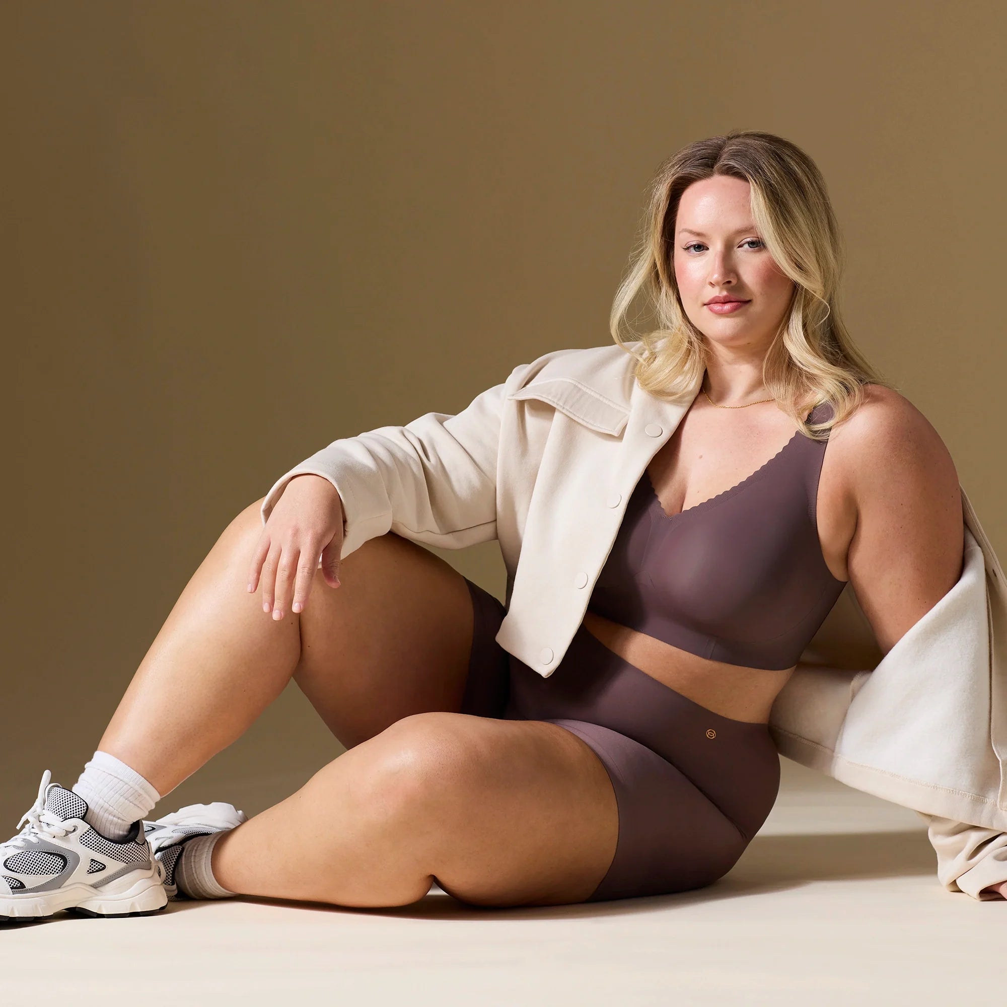 Woman wearing Evelyn & Bobbie Evelyn Bra in Thistle, dark mauve no wire pullover bra, against an olive background, with a coat overtop. 