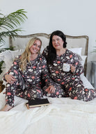 Two women in matching pajamas sitting on a bed with a plant in the background