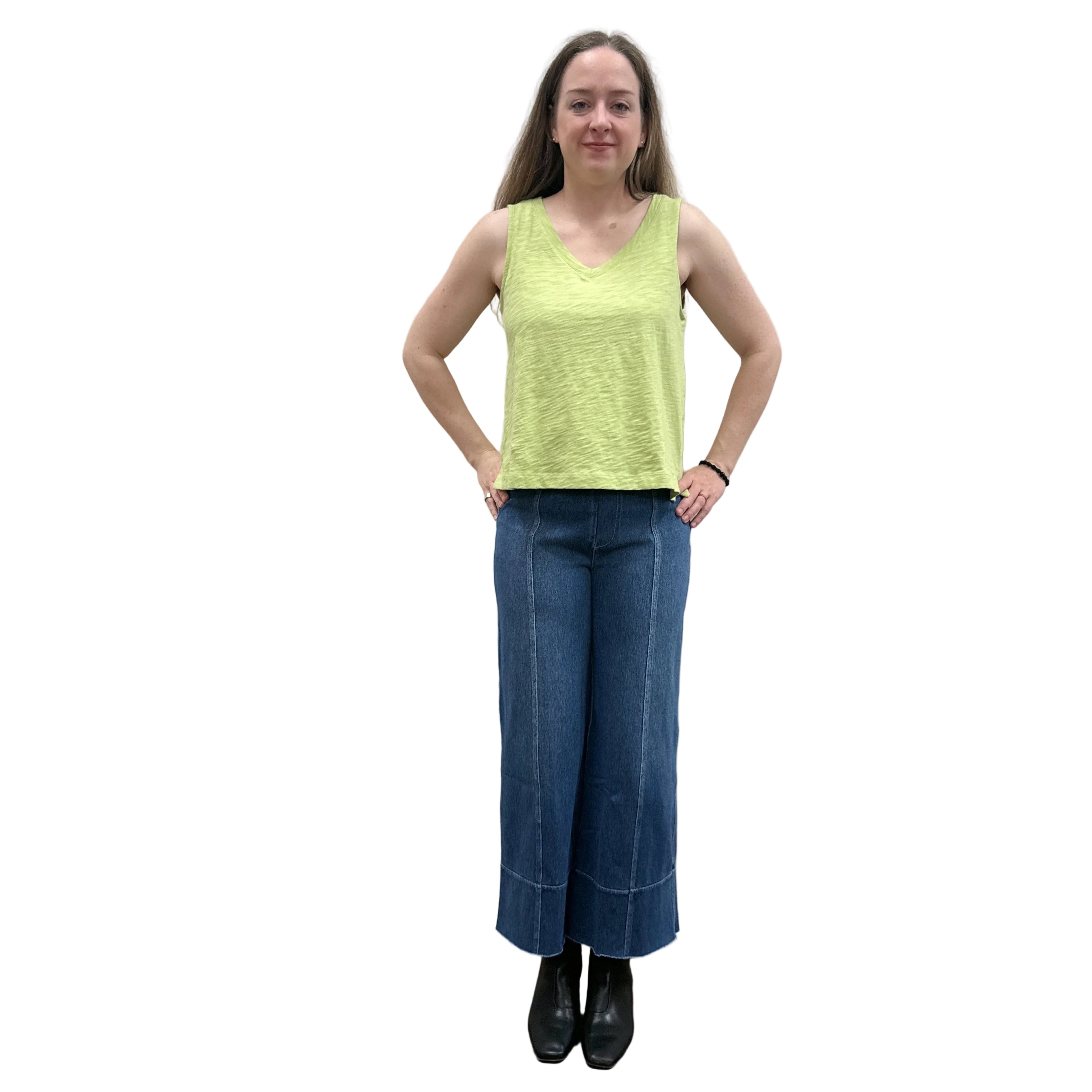 Woman wearing a green sleeveless Papa V-Neck Tank top and blue jeans standing against a plain wall.