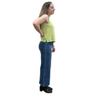 Woman wearing a green sleeveless Papa V-Neck Tank top and blue jeans standing against a plain wall.