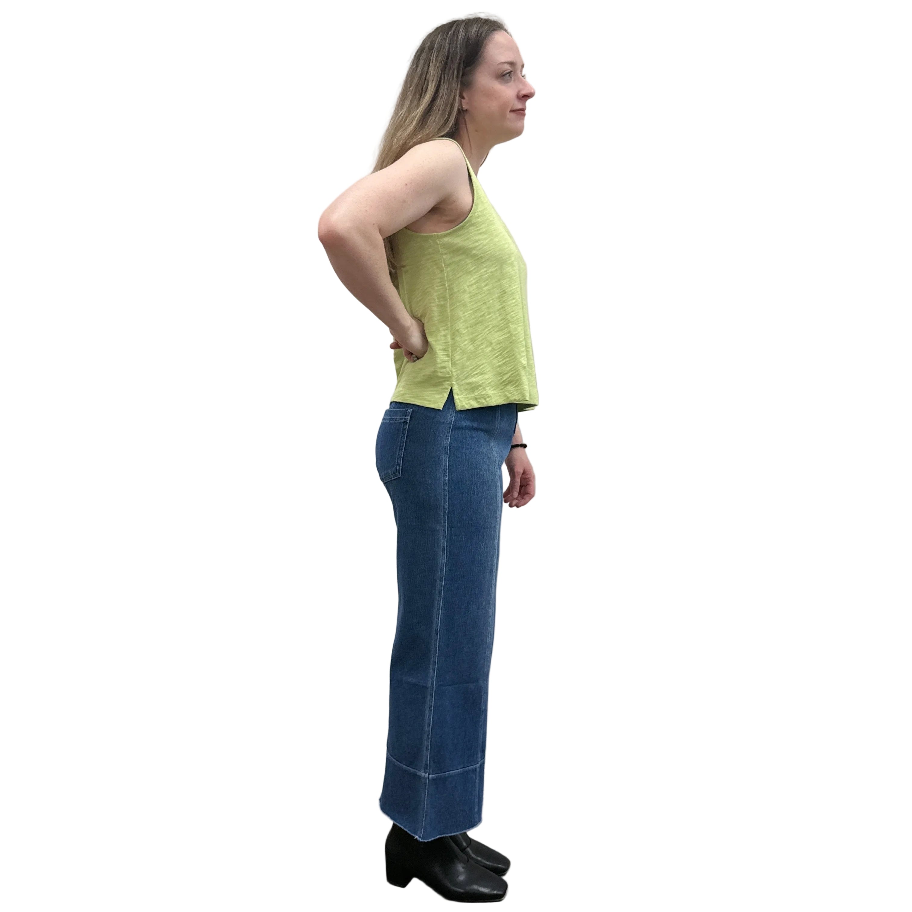 Woman wearing a green sleeveless Papa V-Neck Tank top and blue jeans standing against a plain wall.