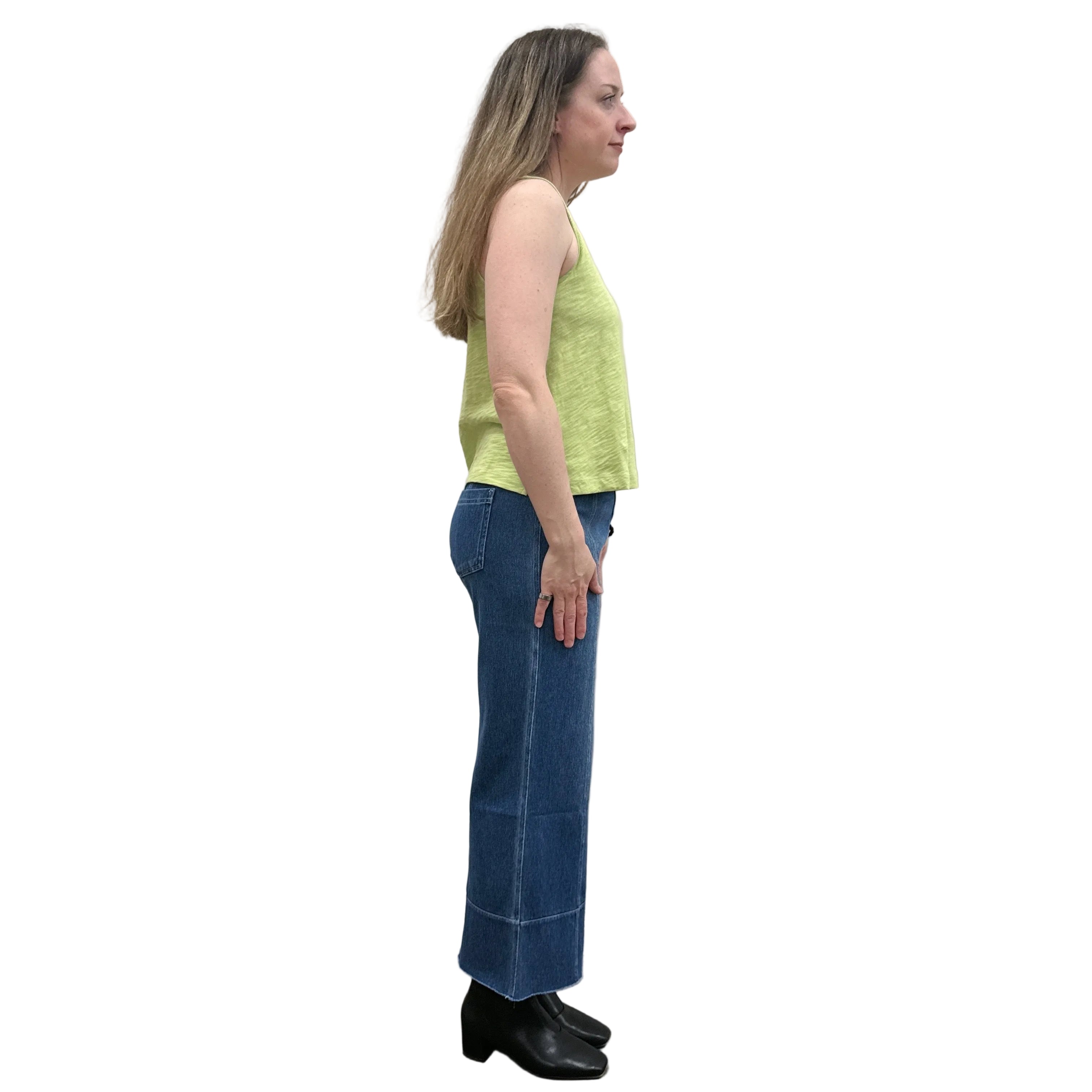 Woman wearing a green sleeveless Papa V-Neck Tank top and blue jeans standing against a plain wall.