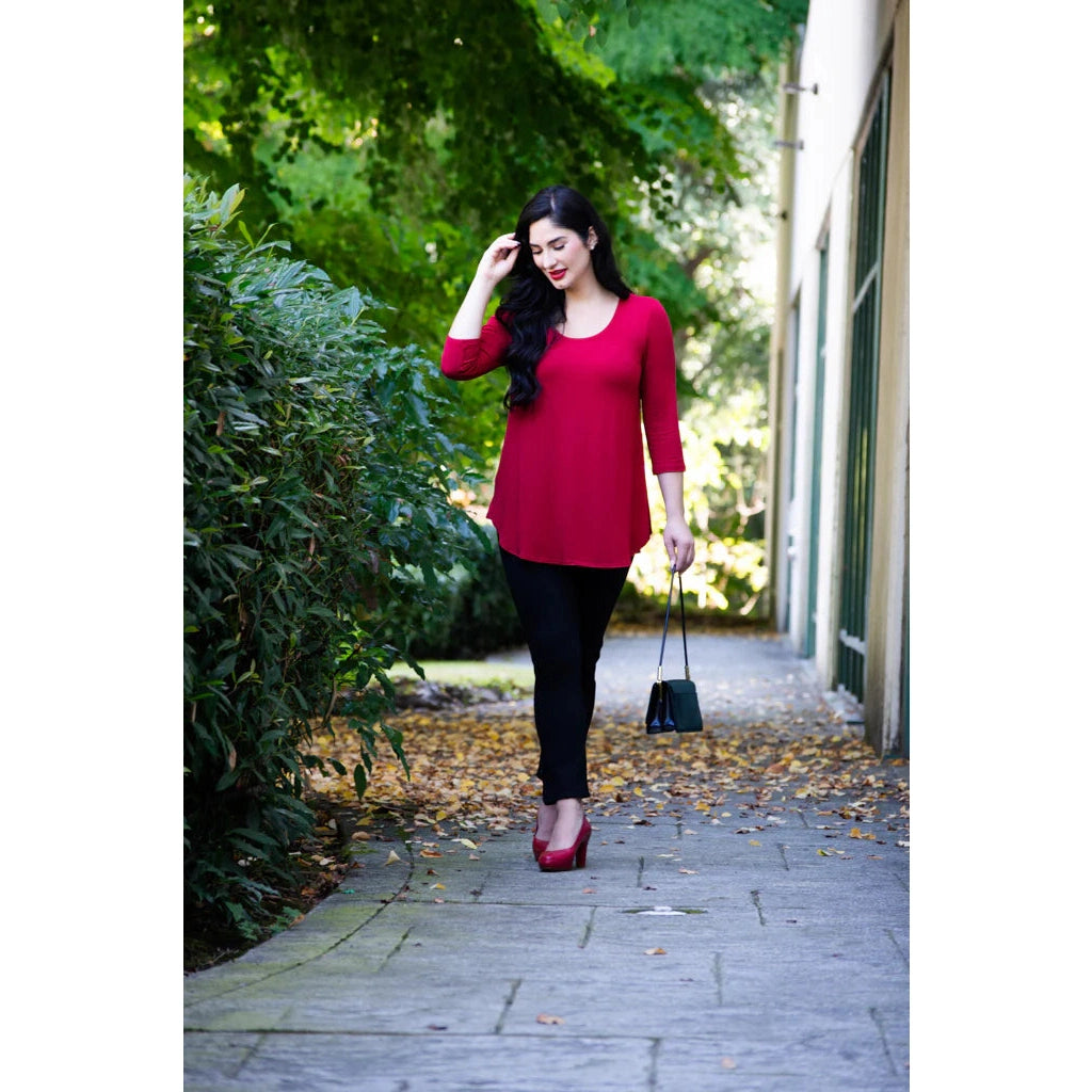 Blue Sky Jazz 3/4 Sleeve Top in Lipstick Red shown on a model wearing black leggings against a greenery background, from the front view. 