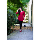 Blue Sky Jazz 3/4 Sleeve Top in Lipstick Red shown on a model wearing black leggings against a greenery background, from the front view. 