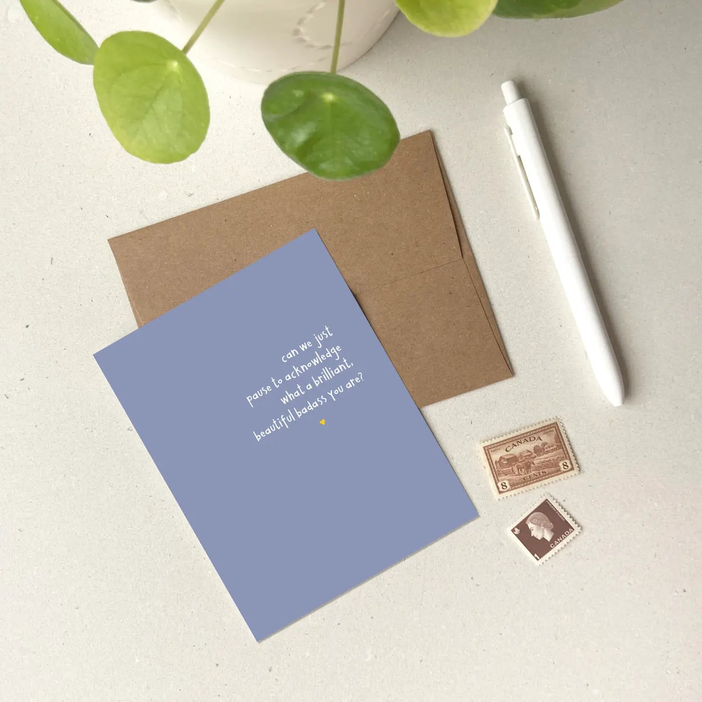 Blue card with text against a beige background with a brown envelope, a pen and stamps. 